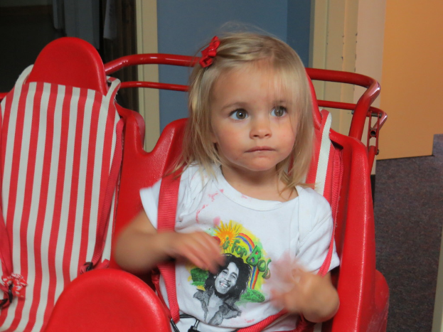 Ein Mädchen in einem weißen T-Shirt sitzt in einem roten Spielzeugfahrzeug an einer Wand.