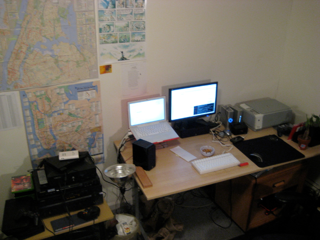 Ein Raum mit einem Tisch, der einen Laptop, Lautsprecher, Monitor, Tastatur, Ball, Maus, Scanner und Papiere enthält, und einem weiteren Tisch mit elektronischen Geräten im Hintergrund, mit Diagrammen und Postern an der Wand dahinter.