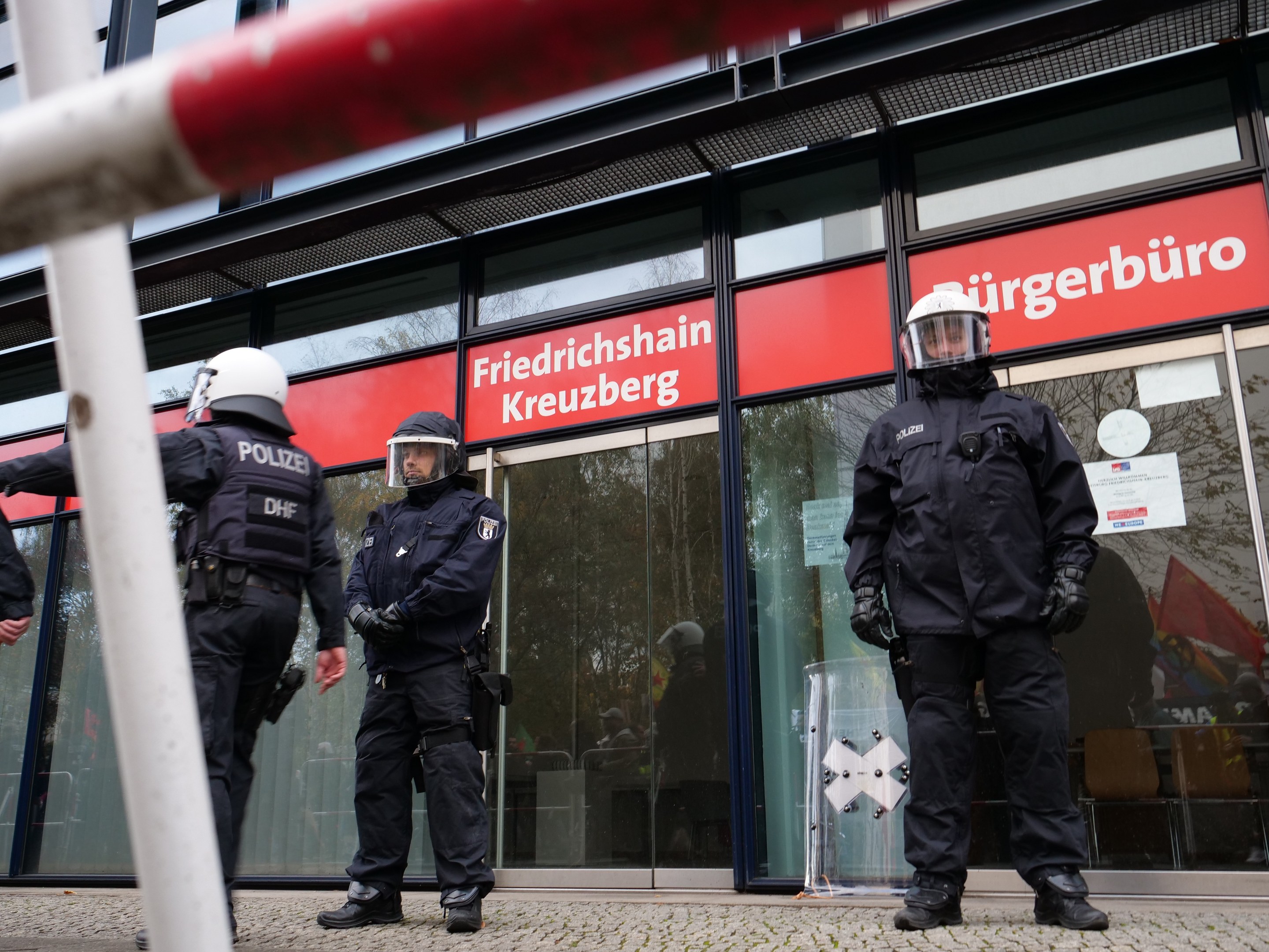 Polizeibeamte in Uniform vor einem Glasfront-Gebäude, einige mit Helmen und Handschuhen, mit einem Pfahl links und Schildern mit "Friedrichshain Kreuzberg" im Hintergrund.