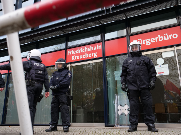 Polizeibeamte in Uniform vor einem Glasfront-Gebäude, einige mit Helmen und Handschuhen, mit einem Pfahl links und Schildern mit "Friedrichshain Kreuzberg" im Hintergrund.