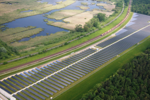 Luftaufnahme eines Solarparks mit Solarpanelen, einem Zug auf einer Eisenbahnschiene, umgeben von Bäumen, Gras, Pflanzen und Wasser.