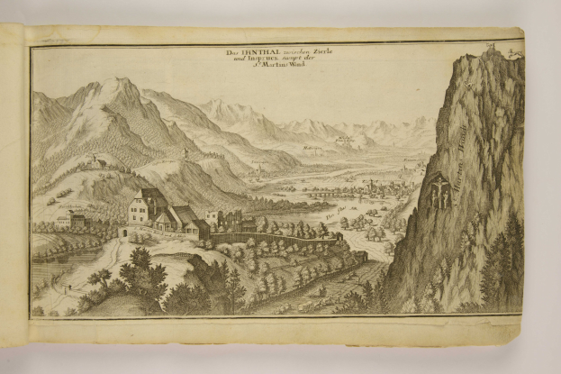 Ein aufgeschlagenes Buch mit einer detaillierten Illustration einer bergigen Landschaft, einschließlich grüner Bäume, verstreuten Häusern, hohen Hügeln und beschreibendem Text.