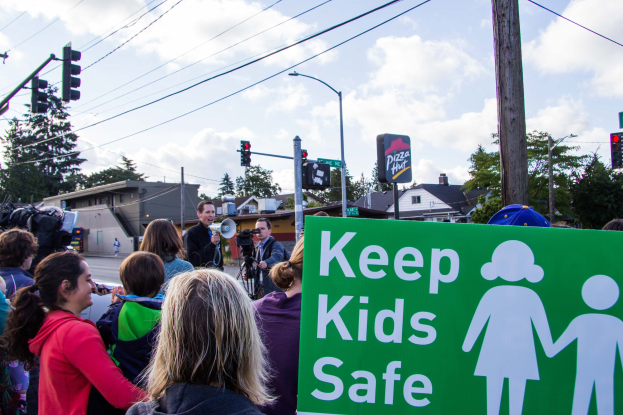 Gruppe von Menschen an einer Straßenkreuzung mit einem "Kinder sicher"-Schild und einem Mann mit Megafon im Hintergrund, mit Gebäuden, Bäumen, Verkehrsschildern, Laternen und Stromleitungen unter einem bewölkten Himmel.