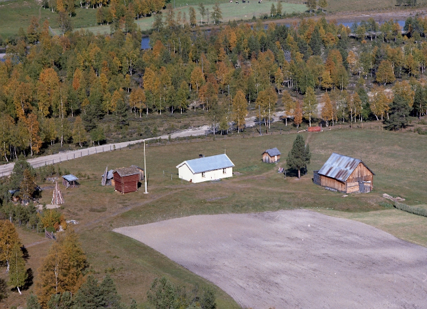 Eine Luftaufnahme eines Ranch mit verstreuten Gebäuden, umgeben von Bäumen, Häusern, Pfählen, einer Straße, Gras und Wasser.