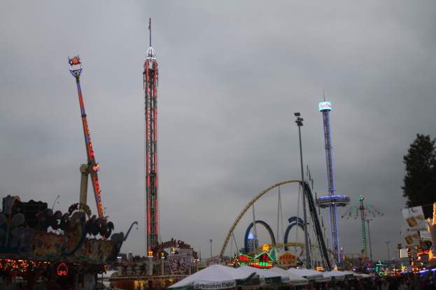 Eine Menschenmenge versammelt sich um einen nächtlichen Fasching, beleuchtet von den Lichtern der Attraktionen, Pfähle und Bäume, unter einem bewölkten Himmel.