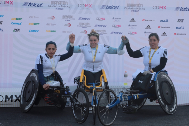 Drei Frauen im Rollstuhl auf einem Podium, die weiße und schwarze Kleider tragen und Medaillen um den Hals haben, heben die Hände zum Sieg.