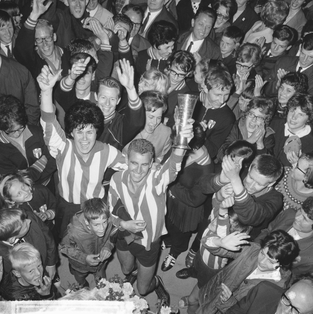 Eine Gruppe von Menschen in Schwarz-Weiß feiert mit einem Pokal vor einer Menge, mit Blumen am unteren Bildrand.