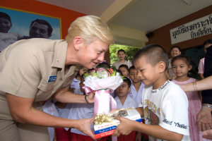 Eine Frau in Uniform überreicht einem jungen Jungen eine Geschenkbox, der einen Blumenstrauß hält, vor einer Gruppe von Kindern und einigen stehenden Personen, einer Tafel mit Schrift auf der rechten Seite und einem Baum im Hintergrund.