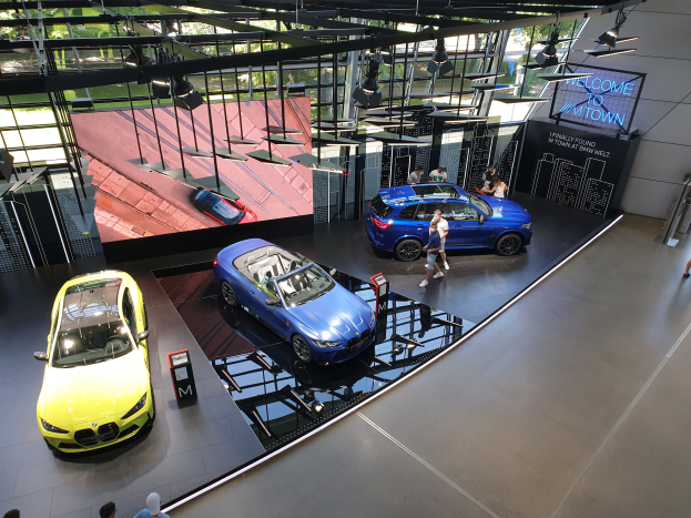 Eine Gruppe von Menschen steht im BMW Museum in München, mit Autos zur Linken und einem Schild mit der Aufschrift "BMW Museum München" zur Rechten, mit Blick auf eine Außenansicht von Bäumen, Gebäuden und Himmel durch Glasfenster.