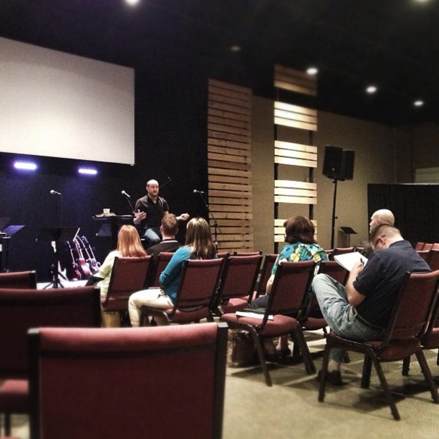 In einer Halle sitzen mehrere Frauen auf roten Ledersesseln, während ein Mann in einem schwarzen Hemd vor ihnen spricht. Hinter ihm befindet sich ein weißer Projektionsschirm, hölzerne Wanddekorationen und ein Lautsprecher auf einem Ständer. Die Decke ist schwarz mit Scheinwerfern.