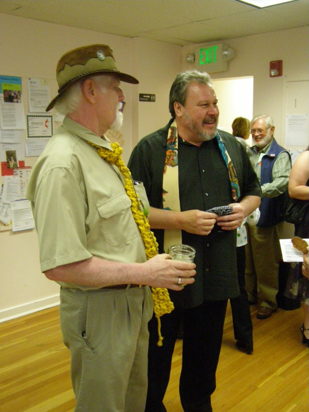 Eine Gruppe älterer Männer, die auf einem Holzboden in einem Raum mit Papieren an den Wänden, einer Warntafel an der rechten Wand, einer Tür darunter und Deckenlampen stehen.