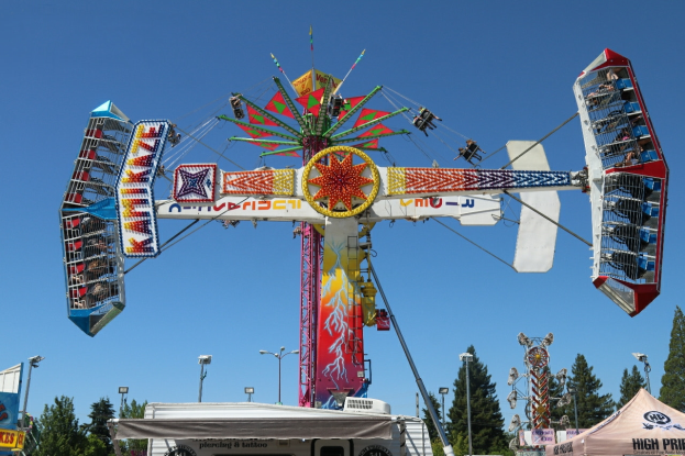 Eine Karussellfahrt auf einem Volksfest mit Menschen darauf, umgeben von Zelten, Bannern, Pfählen, Lichtern, Bäumen und anderen Gegenständen, mit dem Himmel im Hintergrund.