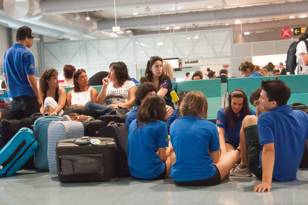 Ein belebter Flughafen-Terminal mit vielen Menschen, die auf Flüge warten, Gepäck links und Glastüren mit Richtungsschildern im Hintergrund.