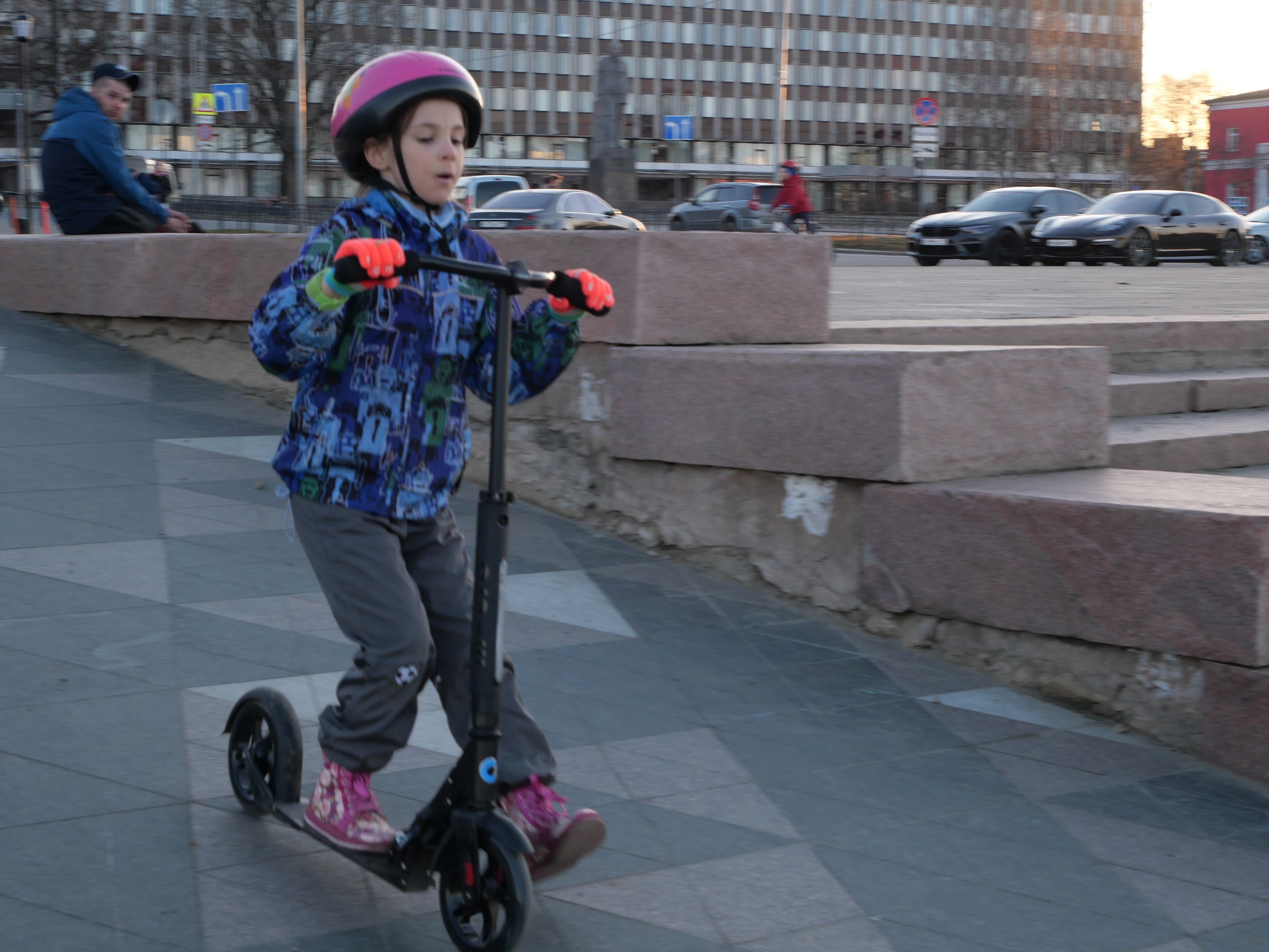 Ein junger Junge in Helm und Handschuhen fährt auf einem Scooter eine Treppe hinunter, Autos, Menschen, Bäume, Pfosten, Bretter, Gebäude und einen klaren blauen Himmel im Hintergrund.