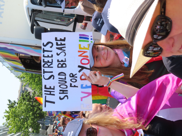 Eine Gruppe von Menschen steht vor einem Lastwagen und hält ein Schild mit der Aufschrift "Die Straßen sollten sicher sein". Einige tragen Mützen und Schutzbrillen, andere halten Schilder. Im Hintergrund sind Bäume, Gebäude und ein klarer blauer Himmel zu sehen.