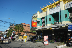 Eine Straßenszene mit einem McDonald's-Restaurant an der Ecke, umgeben von Gebäuden, Bäumen, Straßenlaternen, Strommasten, Stromkabeln, Kraftfahrzeugen, Fußgängern, Werbetafeln, Topfpflanzen und einem klaren blauen Himmel.