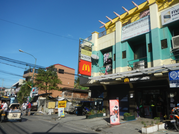 Eine Straßenszene mit einem McDonald's-Restaurant an der Ecke, umgeben von Gebäuden, Bäumen, Straßenlaternen, Strommasten, Stromkabeln, Kraftfahrzeugen, Fußgängern, Werbetafeln, Topfpflanzen und einem klaren blauen Himmel.