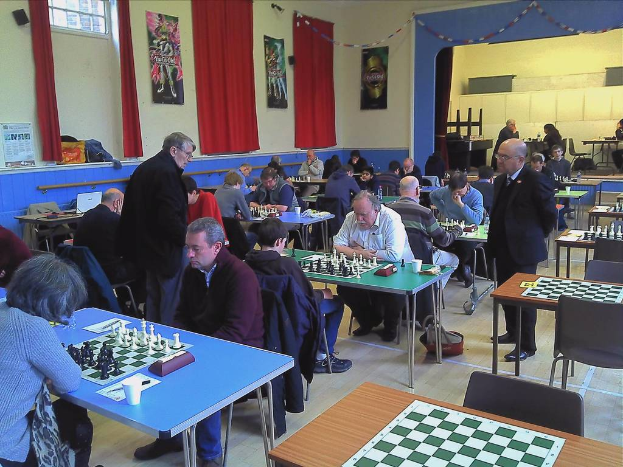Zwei Personen spielen Schach an einem Tisch mit Münzen in einem großen Saal, mit Vorhängen, Plakaten, einem Fenster und einem Kalender an der Wand.