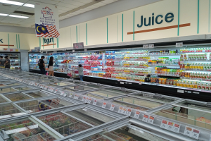 Ein hell erleuchteter Supermarktwöng mit Regalen voller Flaschen, verpackten Lebensmitteln und frischem Gemüse, einige Einkäufer und Deckenbeleuchtung.