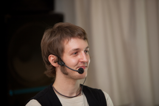 Ein Mann in einem cremefarbenen T-Shirt hält ein Mikrofon vor einem Vorhang, möglicherweise mit Lautsprechern auf der linken Seite.