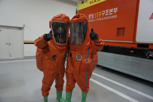 Zwei Personen in orangefarbenen Chemikalienschutzanzügen stehen nebeneinander vor einem Truck, mit einer Wand und Türen im Hintergrund.