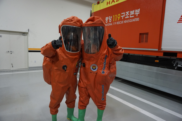 Zwei Personen in orangefarbenen Chemikalienschutzanzügen stehen nebeneinander vor einem Truck, mit einer Wand und Türen im Hintergrund.