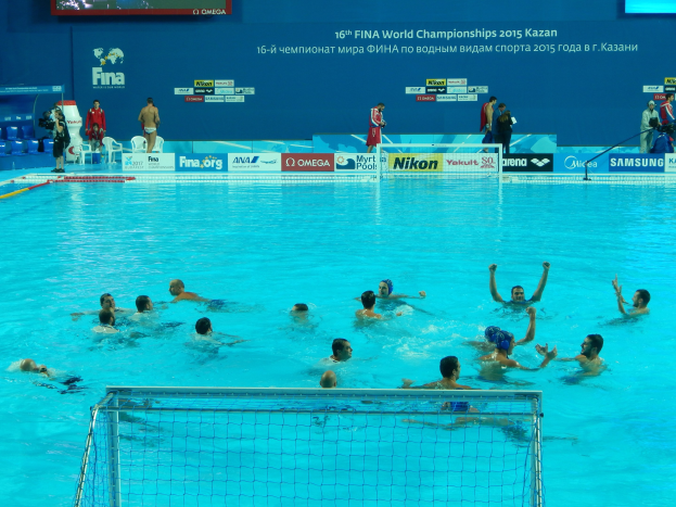 Water polo players competing in a pool during the FINA World Championships 2015 Kazan, with spectators and scoreboards in the background.