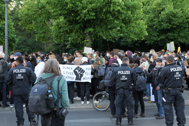 Eine große Gruppe von Menschen, die auf der Straße stehen, einige mit Schildern, mit einem Fahrrad im Vordergrund und Bäumen und einem Pfahl im Hintergrund, bei einer Black-Lives-Matter-Demonstration in Berlin dabei.