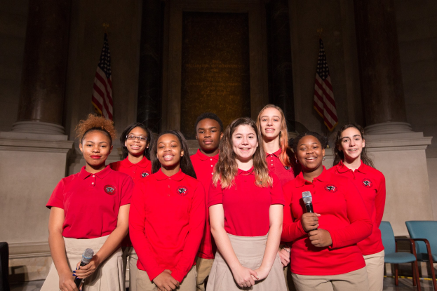Gruppe junger Frauen in roten Shirts, die strahlend Mikrofone halten und vor Stühlen, Säulen und Flaggen stehen, wahrscheinlich Teil eines nationalen Jugendchors.