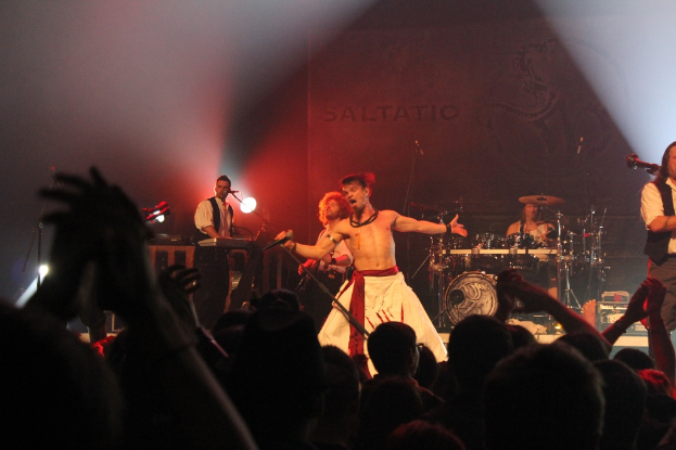 Drei Menschen, die Musikinstrumente spielen, wobei ein Mann ein Mikrofon hält und singt, unter einem fokussierten Scheinwerferlicht, vor einem Publikum.