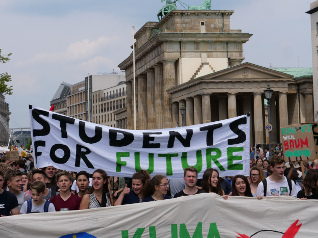 Gruppe von Schülern marschiert in Berlin mit einer bunt bemalten "Students for Future"-Schultertasche vor Gebäuden, Bäumen und Himmel.