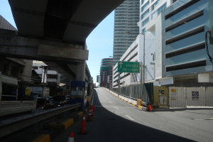 Stadtstraße mit Verkehrsleitkegeln, Fahrzeugen, einer Brücke auf der linken Seite, Gebäuden mit Namensschildern, Laternenpfählen und einem Himmel im Hintergrund.
