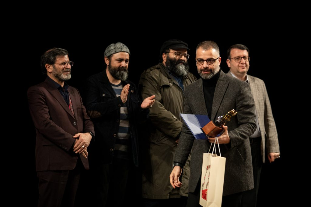 Eine Gruppe von Männern auf der Bühne der Eröffnungsnacht des Berliner Filmfestivals, einer hält eine Tasche und eine Trophäe, mit einem dunklen Hintergrund.