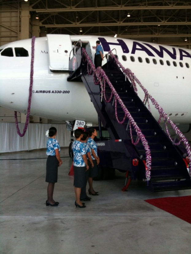 Ein Flugzeug mit Text darauf, eine Treppe, einige stehende Personen, Vorhänge und ein Metallzaun im Hintergrund.