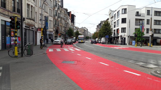 Rote Fahrradspur neben einer Stadtstraße mit fahrenden Fahrzeugen, Fußgängern auf dem Gehweg, Gebäuden mit Fenstern, Bäumen und einem klaren blauen Himmel im Hintergrund.