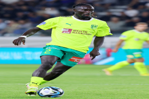 Ein Fußballspieler in gelbem T-Shirt und grünen Shorts ist im Einsatz auf dem Feld, mit einem blauen Board und unscharfen Menschen im Hintergrund.