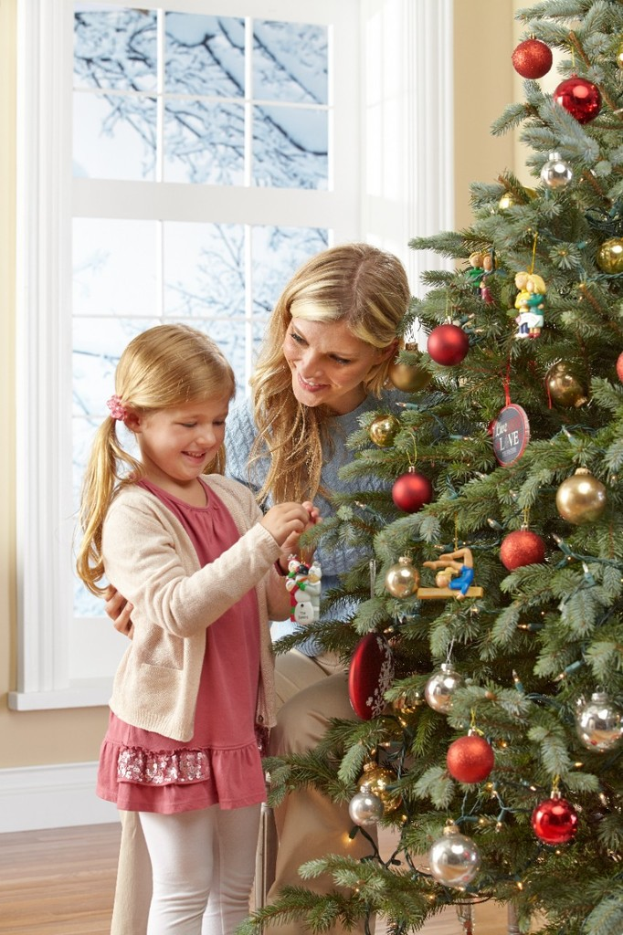 Ein geschmückter Weihnachtsbaum mit einer Frau und einem Kind davor, ein Fenster und eine Wand im Hintergrund und Bäume vor dem Fenster.