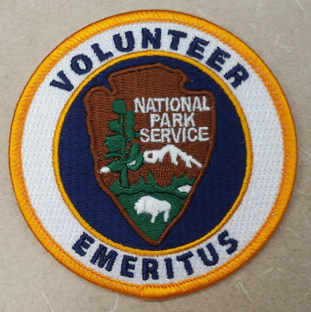 Ehrenamtlicher Nationalparkdienst-Abzeichen mit blauer Umrandung, weißem Hintergrund und gelbem Kreis um die zentrierte schwarze Schrift.