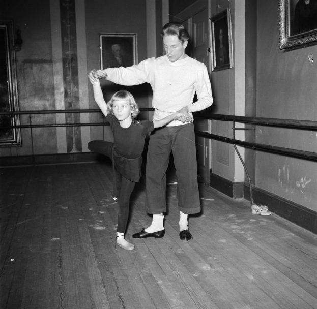 Ein Mann in weißem Hemd und schwarzer Hose tanzt mit einem kleinen Mädchen in einem Tanzstudio, mit Fotorahmen an den Wänden und einem Geländer im Hintergrund, alles in Schwarz-Weiß dargestellt.