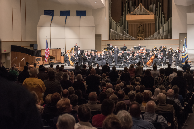 Eine große Gruppe von Menschen sitzt vor einer Bühne mit einigen, die Musikinstrumente halten. Flaggen mit Stangen, ein Lautsprecherbox, andere Gegenstände auf der Bühne, eine Decke, Deckenleuchten und eine Tür an der Rückwand deuten auf ein Konzert an der University of Texas at Austin hin.