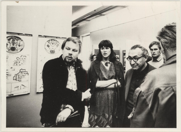 Eine Gruppe von Menschen, die in einem Raum mit einer plakatgeschmückten Wand stehen und sprechen, schwarz-weiß, 1980er Jahre.