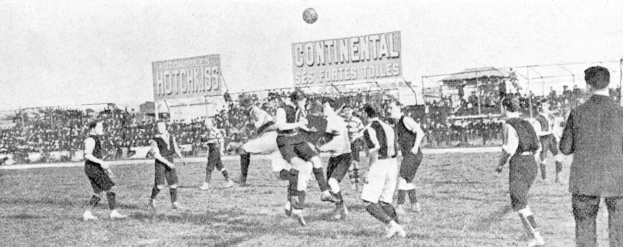 Schwarzes Bild von Männern, die auf einem Feld Fußball spielen, mit einem Ball in der Luft, Zuschauern im Hintergrund und Anzeigetafeln mit Spielinformationen.