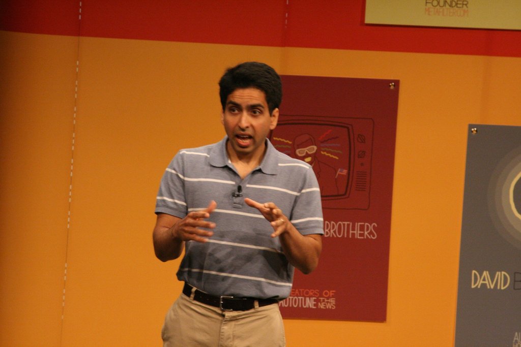 Ein Mann steht auf der Bühne und spricht vor einer Wand mit Plakaten