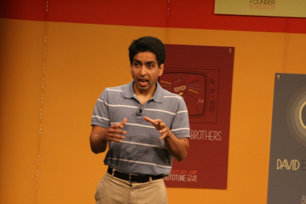 Ein Mann steht auf der Bühne und spricht vor einer Wand mit Plakaten