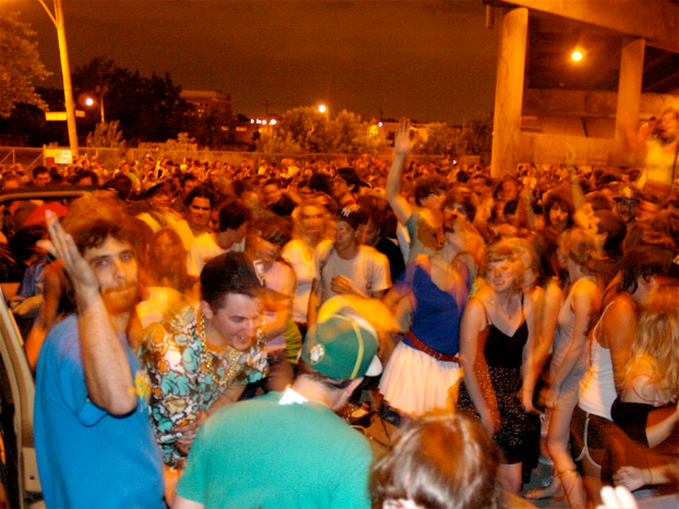 Menschen, die im Vordergrund eines Konzerts mit Lichtern, Pfählen, Bäumen und anderen Gegenständen im Hintergrund tanzen.