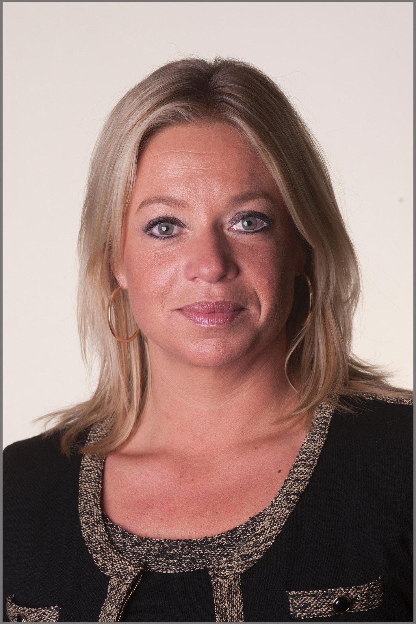 Eine Frau mit blonden Haaren in einem ordentlichen Dutt, die ein schwarzes Kleid und goldene Kreolen trägt und warm und einladend lächelt mit leuchtenden, lebendigen Augen.