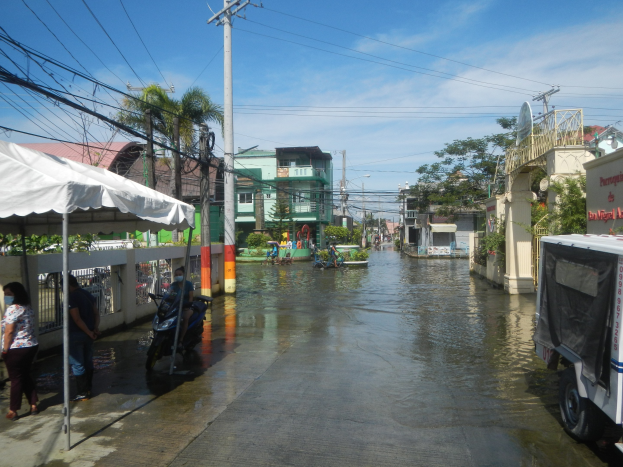 Eine überflutete Stadtstraße mit Fahrzeugen, Fußgängern, einem Zelt, Strommasten, Gebäuden, Bäumen und einem bewölkten Himmel.