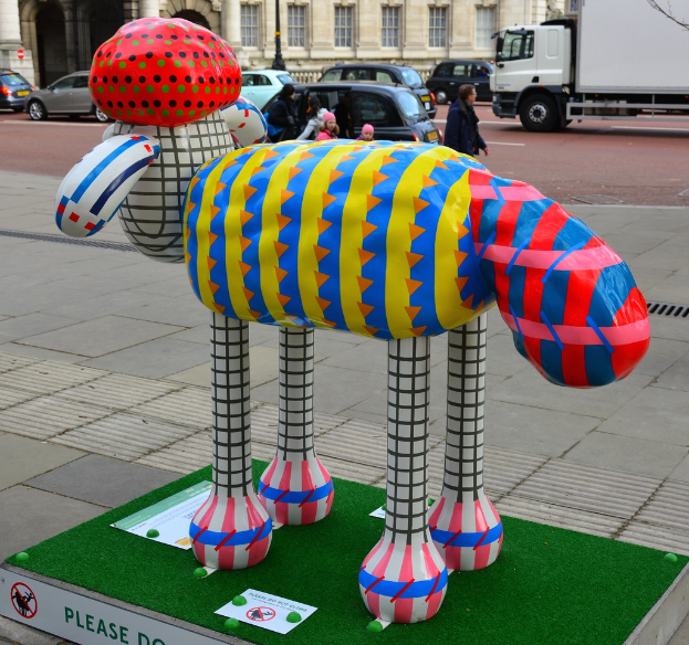 Eine Skulptur auf dem Boden mit Brettern in der Nähe, fahrende Fahrzeuge auf der Straße dahinter, ein paar Menschen auf dem Gehweg und Gebäude mit einem Mast im Hintergrund.