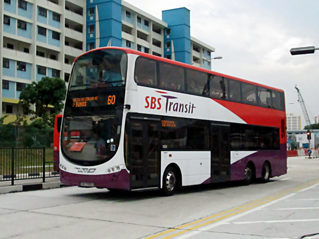 Ein elektrischer Bus steht im Mittelpunkt der Szene mit einem Gebäude im Hintergrund, Bäumen vor dem Gebäude, einem Zaun auf der rechten Seite des Fußwegs und Himmel oben.