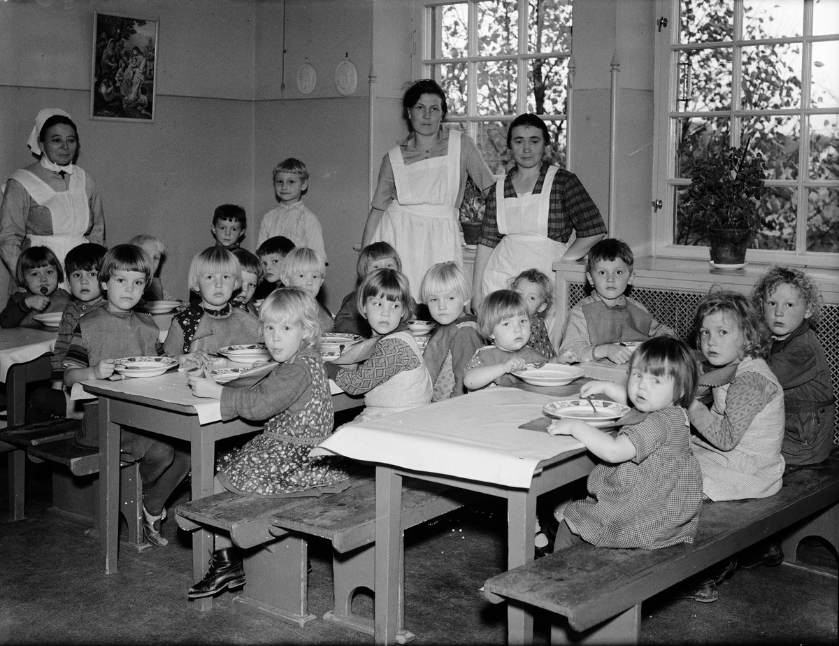 Kinder sitzen an Tischen in einem Klassenzimmer mit Tellern voller Essen, umgeben von Bänken, einem Foto auf der Wand und Fenstern mit Pflanzen draußen.